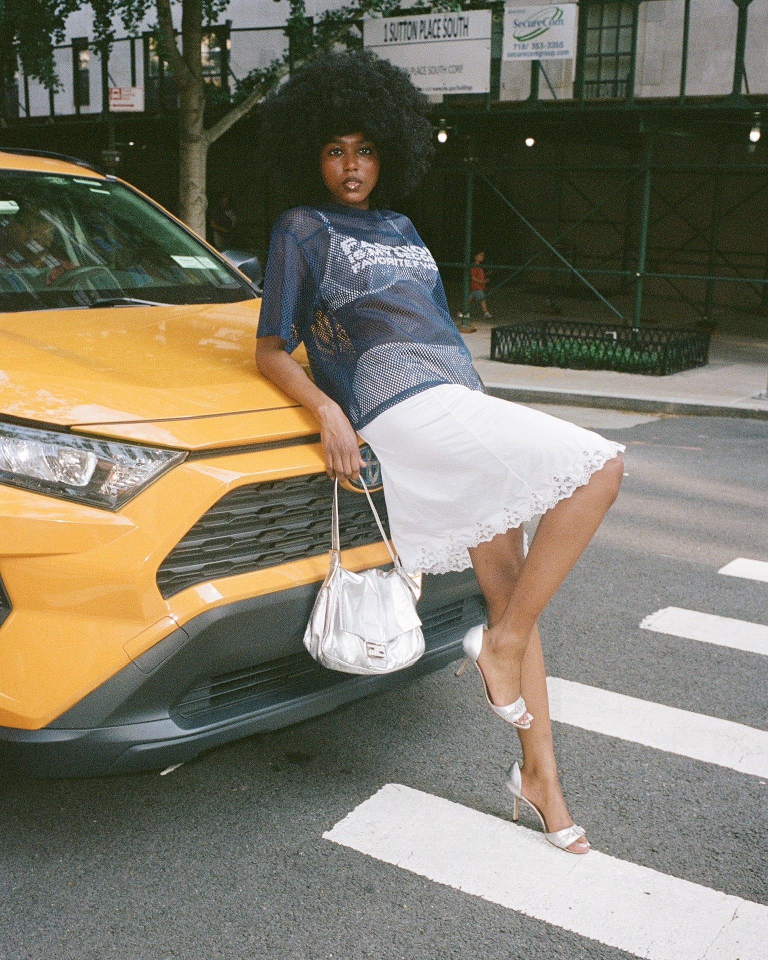 A women leans confidently onto a taxicab. She wears an afro, a mush jersey top, a white skirt and is holding a handbag.